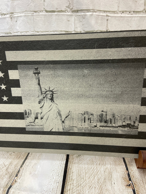 A scene of New York city skyline and the Statue of Liberty on the US flag. All laser engraved onto a piece of black slate.