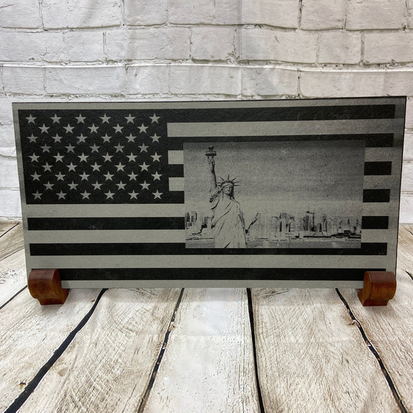 A scene of New York city skyline and the Statue of Liberty on the US flag. All laser engraved onto a piece of black slate.