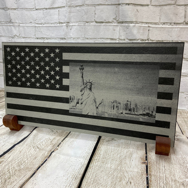 A scene of New York city skyline and the Statue of Liberty on the US flag. All laser engraved onto a piece of black slate.