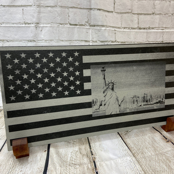 A scene of New York city skyline and the Statue of Liberty on the US flag. All laser engraved onto a piece of black slate.