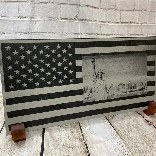 A scene of New York city skyline and the Statue of Liberty on the US flag. All laser engraved onto a piece of black slate.