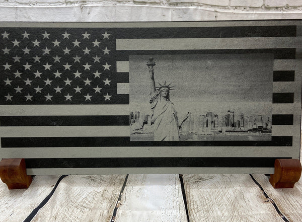 A scene of New York city skyline and the Statue of Liberty on the US flag. All laser engraved onto a piece of black slate.