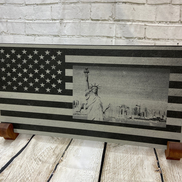 A scene of New York city skyline and the Statue of Liberty on the US flag. All laser engraved onto a piece of black slate.