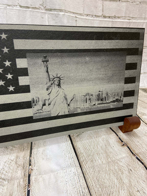 A scene of New York city skyline and the Statue of Liberty on the US flag. All laser engraved onto a piece of black slate.